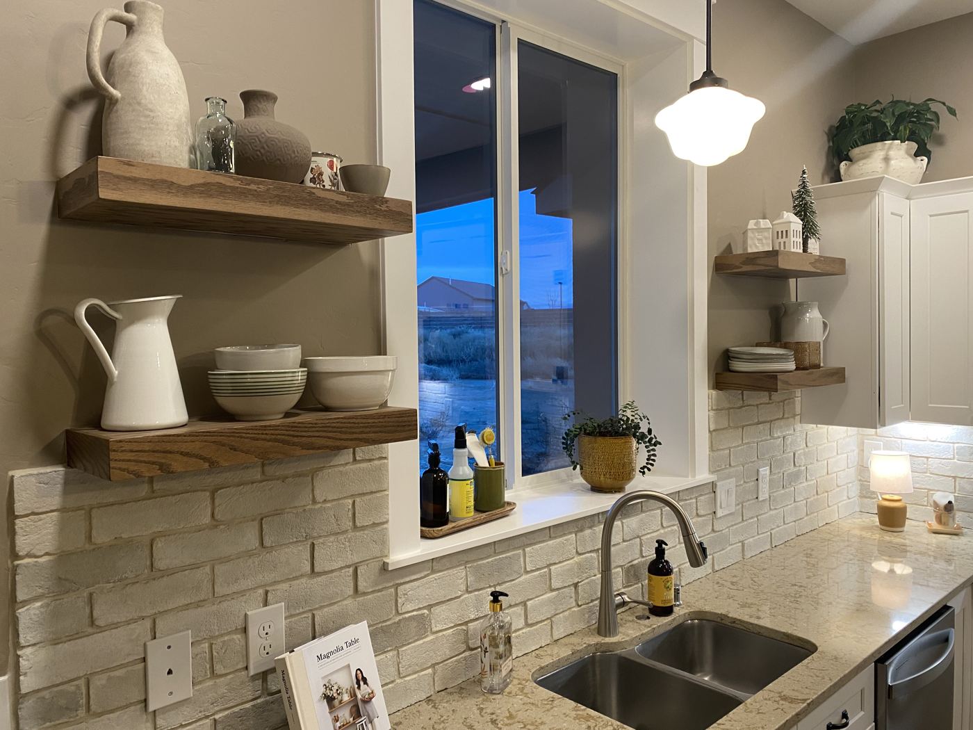 White shaker kitchen with stone counters, brick backsplash and wood shelves